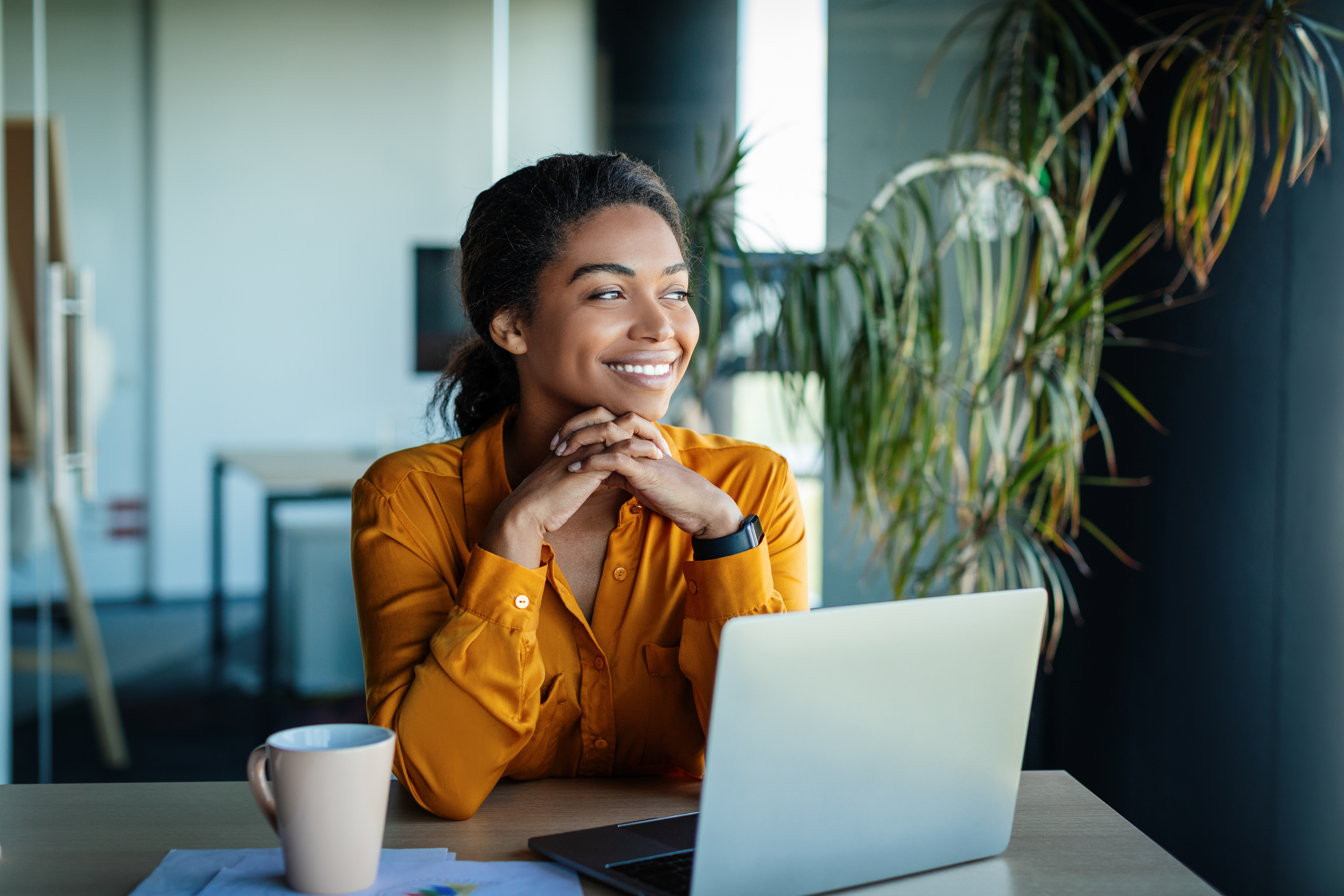 Portrait of dreamy african american businesswoman sitting at desk in office and thinking while working on laptop, looking away with pleased face expression, copy space
