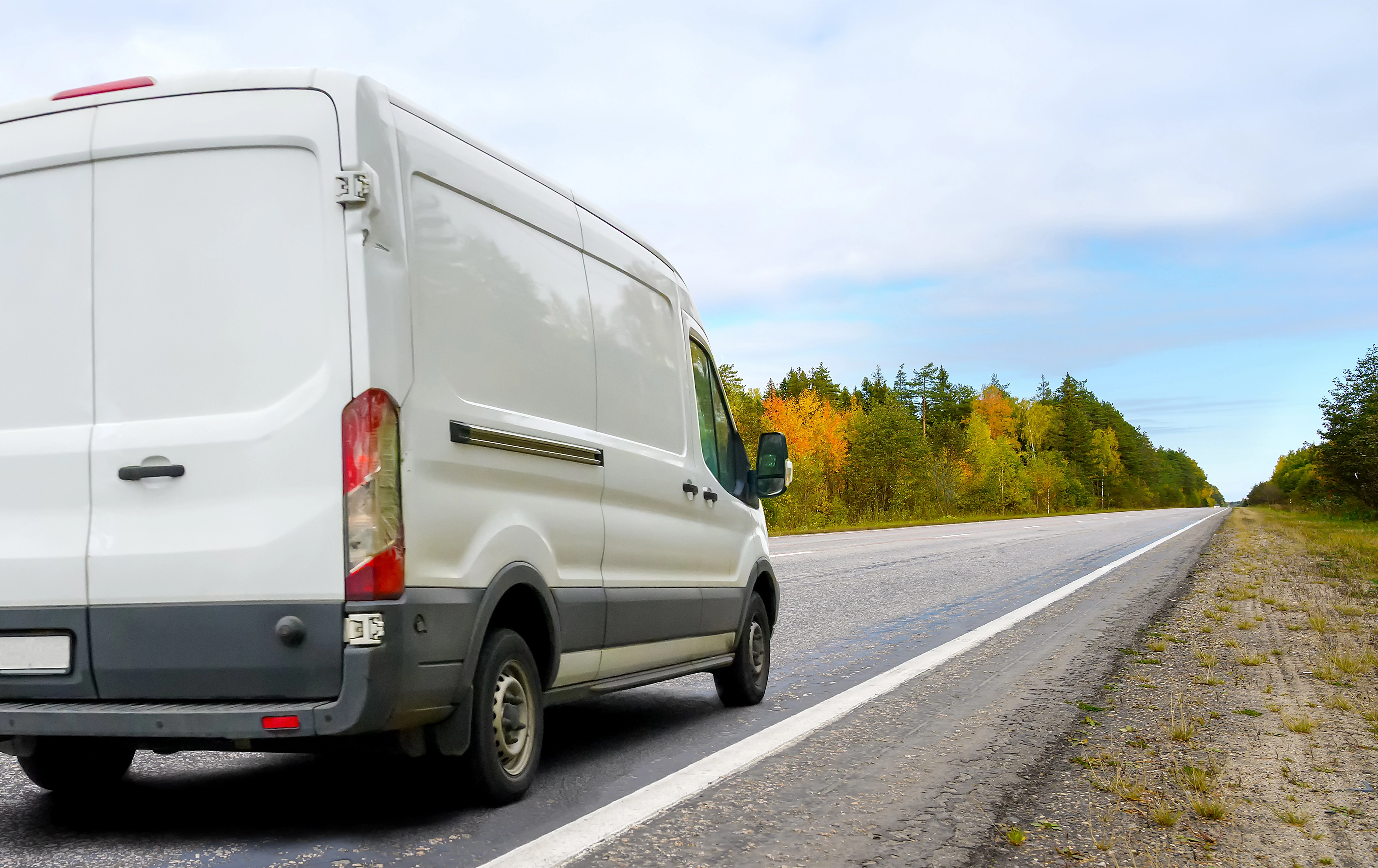 A white cargo van driving down an empty, wooded road